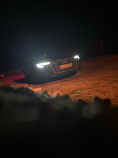 The image displays a dark-colored Audi sedan, likely an A6 model, parked on unpaved ground during nighttime. The vehicle is angled towards the viewer's right, with its bright white LED headlights fully illuminated, casting a strong, warm orange glow on the dirt surface directly ahead. The front grille, featuring the Audi four-ring emblem, is prominent. A European license plate, marked "B AI 2815", is affixed to the lower front bumper. To the left and in the background, a second dark-colored vehicle is partially visible, its rear section and a red illuminated taillight reflecting on the ground. The ground is characterized by orange-brown dirt and sparse vegetation. The background is largely dark, with a slender, dark vertical post visible on the far right. The immediate foreground is blurred and significantly darker, obscuring details. No individuals are visible in the scene. The scene is located in the city of Novgrad, Bulgaria.