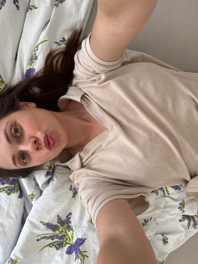 A woman lies on a bed with a white and purple floral-patterned bedsheet. She wears a beige sweater with a V-neck. The woman faces the camera with her mouth slightly puckered, as if taking a selfie. Her brown hair is visible near her head. The location is not discernible from the image. There is nothing to suggest the location is Kőröshegy, Hungary.