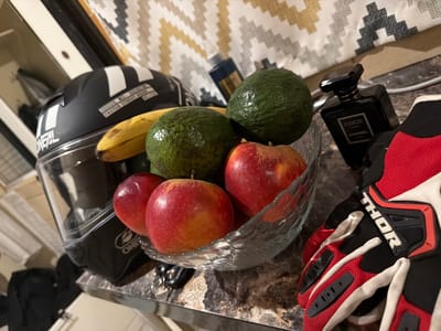 An overhead shot captures a still life arrangement on a speckled brown countertop. In the foreground, a red and black motorcycle glove rests on its side. To its left, a clear glass bowl overflows with fruit, including three red apples, two green avocados, and a partially visible yellow banana. Behind the fruit bowl, a black motorcycle helmet with white stripes is positioned. Further back, a black perfume bottle, labeled "COCO CHANEL," stands upright. The background features a zig-zag patterned fabric in shades of cream, yellow, and grey. This scene is located in Chereshnya, Russia.