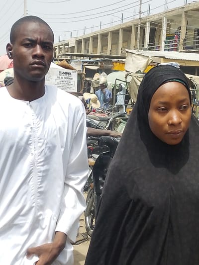 The image depicts an outdoor street scene in Fagge D1, Nigeria, under bright daylight conditions. In the foreground, two individuals are present: a male on the left and a female on the right. The male, with dark skin and short dark hair, wears a white long-sleeved tunic (tobi) with vertical embroidery detail on the chest and a stand-up collar. A white wireless earbud is visible in his right ear. He gazes directly forward, appearing serious. His left hand is partially visible, tucked into his garment. The female, also with dark skin, wears a black hijab covering her head and shoulders, with a hint of a green lining or underscarf visible near her forehead. She has a small nose piercing on her right nostril and her gaze is directed towards the right side of the frame, slightly downward. There is no direct interaction evident between the two foreground subjects.
Behind the individuals, the environment suggests a bustling market or commercial area. A grey and white box prominently features "ABRO MOTORS" and "500 OIL TREATMENT," along with specifications such as "24 CS," "PART No. AB-5030 AM," "24 x 15 FL OZ. (443ML) CANS," and "Aabro Industries, Inc. Made in U.S.A." This box is positioned near what appears to be a stall or goods display. Various market stalls with corrugated metal roofs and assorted merchandise are visible further back. Several other individuals are scattered in the background; one person in a blue garment appears to be carrying a large object on their head. A black motorcycle with chrome accents is partially visible between the two foreground subjects, positioned behind the male and to the left of the female. In the distant background, a multi-story concrete building with numerous open rectangular window or balcony frames indicates ongoing construction or an unfinished structure. Overhead wires are also visible. The ground appears to be unpaved, consistent with a dusty or sandy surface. The overall lighting is harsh, suggesting strong sunlight.
