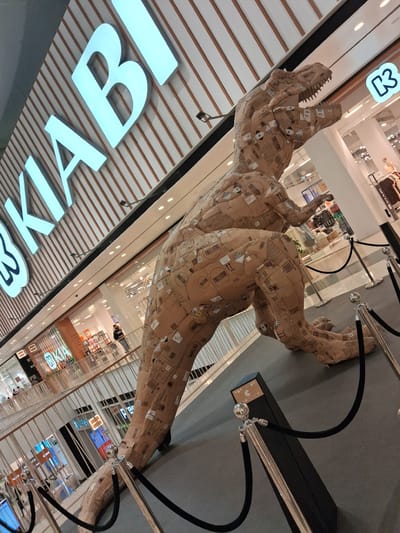 Here is a description of the image:

The image shows a large dinosaur sculpture made from cardboard boxes inside a shopping mall. The sculpture is tan and covered in labels and barcodes. It's surrounded by black rope barriers supported by chrome posts. Below the sculpture is a section of the shopping mall with the “KIABI” store in view. The location is likely in Amadora, Portugal, given the presence of the "KIABI" store. The photo is taken from a low angle, looking up at the sculpture.