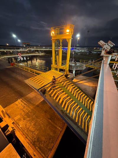 The image depicts a ferry ramp connected to a ferry at a port at night. A person is seen crouching on the ferry ramp. The ramp is yellow and green with diagonal black stripes. The ferry is docked next to a pier with yellow railings and lights. In the background, there are buildings, lights, and a dark, cloudy sky. The location is identified as Melaya, Indonesia.