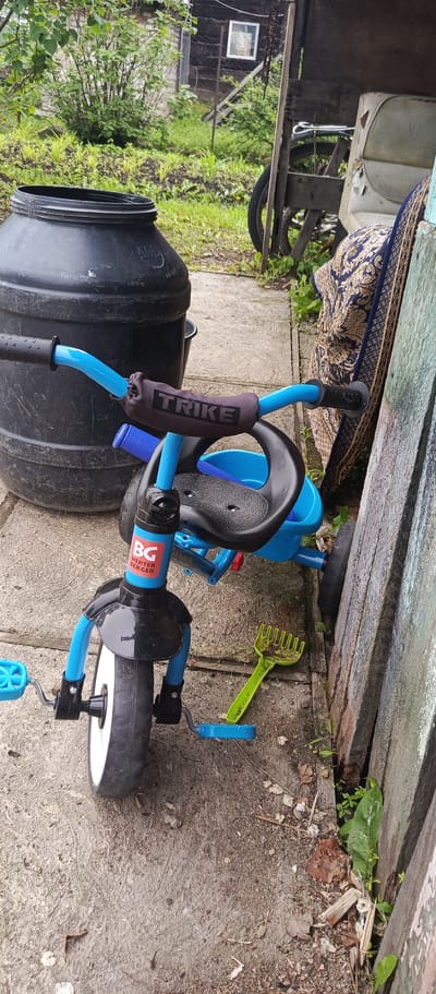 A blue and black child's tricycle is parked on a concrete surface next to a large black plastic barrel and a wooden fence. The tricycle has "TRIKE" printed on a dark grey cover on its handlebars. A small green plastic rake lies on the ground near the tricycle. In the background, a dilapidated wooden structure with a small window is visible, surrounded by greenery. The scene appears to be an outdoor area, possibly a yard or storage space in the city of Pogranichny, Russia.