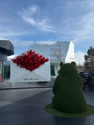 The scene depicts an outdoor area at The Village outlet in La Verpillière, France, under a bright blue sky with scattered cirrus clouds. In the right foreground, a large, dark green topiary shaped as a sitting bear faces away from the camera, resting on a circular patch of artificial turf. Centrally in the midground, a prominent white sculpture resembling an open envelope is filled with a dense cluster of red artificial flowers. The text "THE VILLAGE" is printed in black capital letters on the envelope's lower flap. This installation is positioned against a multi-paneled mirrored wall, which reflects distorted images of a bare tree, the sky, and faint human figures walking past. To the far right, beyond dark barriers, a parking lot with vehicles and several trees is visible. The ground is a dark, textured pavement with shadows cast from left to right.