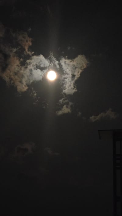 The image depicts a nighttime scene with a bright full moon partially obscured by clouds. The moon is located in the upper center of the frame. The sky is dark, with some wispy clouds illuminated by the moonlight. In the lower right corner, the edge of a man-made structure, possibly a building or lamp post, is visible against the dark sky. The image does not provide any specific visual cues or context that would allow for the identification of its location in Viterbo, Italy.
