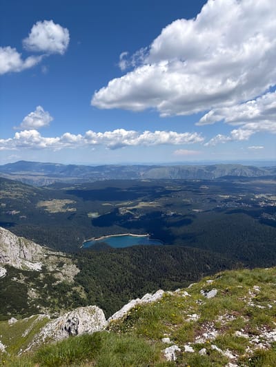 This is a panoramic, high-angle shot of a mountainous landscape, likely taken from a peak or high vantage point. The sky is a vibrant blue, dotted with fluffy white cumulus clouds. In the mid-ground, a dense, dark green coniferous forest stretches across rolling hills, with a striking, turquoise lake nestled within it. Beyond the forest, a vast expanse of layered mountains recedes into the distance, their peaks softened by atmospheric haze. The foreground features rocky, uneven terrain covered in patches of green grass and small wildflowers, indicating a natural, undeveloped environment. The scene is devoid of any human presence or man-made structures, suggesting a remote, natural setting within the Durmitor National Park, near Motički gaj, Montenegro.