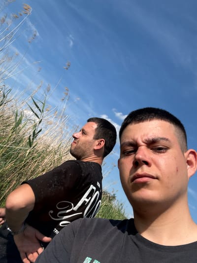 The image features two men outdoors on a clear, sunny day in Zamfir, Bulgaria. The photograph is taken from a low angle, looking upwards towards a bright blue sky with sparse, wispy white clouds.

In the foreground, an adult male with short dark hair and light stubble looks directly towards the camera with a serious, intense expression. He has a slight furrow in his brow and a small red mark is visible on his forehead. He is wearing a plain black t-shirt.

Behind him, slightly to his right, stands a second adult male, shown in profile. He has dark hair and a beard, and appears to be looking downwards or away, possibly in contemplation or focus. He is also wearing a black t-shirt. Visible white text on his shirt includes "FO[R/Y]OU" prominently, along with partial letters like "EST" and "TR". His hands are partially visible, held together at waist level.

The lower half of the image and extending into the mid-ground is filled with tall, dry reeds or grass, suggesting a natural, possibly wetland or rural environment. Bright sunlight casts strong shadows on the subjects, particularly on the neck and jawline of the foreground man.