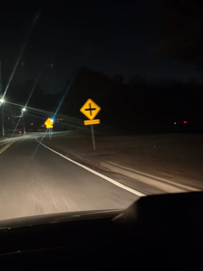 A dark and blurry image depicts a road at night. A yellow diamond-shaped road sign with a black cross inside, indicating a crossroad, is visible on the left side of the road. Another similar sign appears smaller further down the road. The road has a white line marking the edge. The right side is largely dark, showing part of a vehicle's interior.