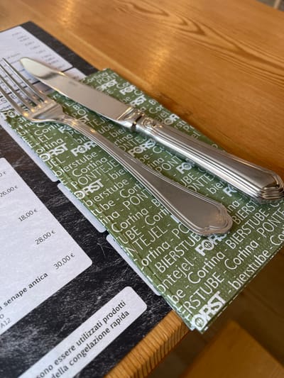 This image shows a close-up of a table setting at a restaurant. The subjects are a fork and knife resting on a green paper napkin with white text. The napkin has repeated patterns of text including "ORST", "BIERSTUBE", "Cortina", and "PONTEJE". Below the napkin is a menu with various prices, including "26,00 €", "18,00 €", "28,00 €", and "30,00 €". There is also text in Italian on the menu and a separate white slip, which reads "sono essere utilizzati prodotti della congelazione rapida" and "senape antica". The background is a light brown wooden table with visible grain. The lighting suggests it is daytime. The presence of cutlery and menus indicates a meal is about to be served or has just concluded. The text "Cortina" on the napkin suggests the location is Cortina d'Ampezzo, a ski resort town in Italy.