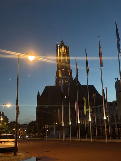 An exterior, eye-level, medium shot captures the Grote Kerk in Arnhem, Netherlands at dusk. The church, a large, dark stone structure with a tall, illuminated tower, dominates the scene. A clock face is visible on the tower. In front of the church, a line of flagpoles with various flags, some illuminated from below, stands prominently. Several flags are visible, including the Dutch flag. To the left of the frame, a streetlamp with a bright orange glow illuminates the foreground. A white Audi car with a license plate reading "JTK-91-R" is parked on the left side of the street. The street is paved with brick and some wet patches are visible on the ground. In the background, other buildings and trees are dimly lit. The sky is a deep blue, transitioning to a lighter hue near the horizon. The overall atmosphere is serene and slightly dramatic due to the lighting.