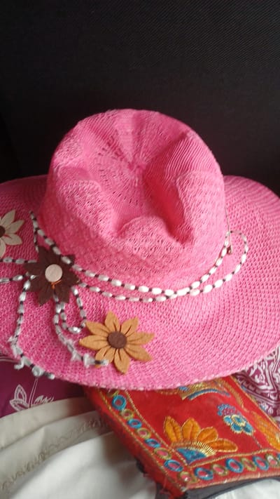 A bright pink woven sun hat with a wide brim is lying on top of a folded red fabric item with embroidered floral designs. The hat is decorated with two felt flowers, one light brown and one tan, each with a brown center. A beaded string with white and black beads, along with some white fringe, is wrapped around the hat's crown and attached to the flowers. The background is dark and textured, appearing to be a dark fabric. The bottom right portion of the image shows a patterned fabric, possibly a blanket or tablecloth, with white and maroon designs. There are no people or visible actions taking place in the image. The scene is likely indoors, given the lighting and background. The location of this image in the city of Киясово, Russia, is not visually discernible from the image itself.