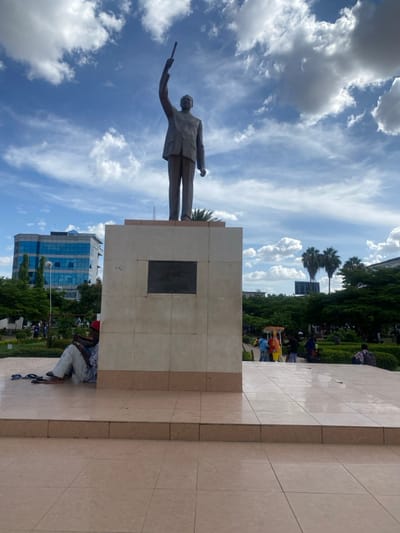 A large bronze statue of a man stands prominently on a tall, light-colored stone pedestal in an outdoor public space in Ihumwa, Tanzania. The man in the statue is depicted in a commanding or dynamic pose, holding a long, slender object resembling a baton or staff in his raised left hand, while his right hand is clenched at his side. He wears a suit-like outfit.
The pedestal, constructed from light-toned, possibly tiled, blocks, features a dark, rectangular plaque on its side, but any inscription on it is illegible due to the image resolution. At the base of the pedestal, to the right, a person wearing light-colored attire and a red cap is seated on the ground, with sandals placed nearby.
The scene is set under a bright blue sky dotted with scattered white, cumulus, and wispy cirrus clouds, indicating clear, sunny weather, likely midday or early afternoon. In the background, modern multi-story buildings with blue-tinted windows are visible, along with abundant green foliage, including palm trees and other deciduous trees, suggesting an urban park or city square environment. Further in the background, colorful, possibly yellow and red, structures are discernable amidst the greenery, with several other individuals scattered throughout the park area. The overall impression is that of a public monument within a vibrant urban landscape.