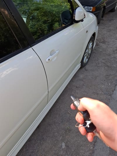 A first-person perspective captures a hand holding car keys near a white car. The keys, featuring a black fob and a metallic keychain, are positioned in the foreground. The white car, with its door handle visible, is parked on a dirt road. In the background, another dark-colored car is visible. The surrounding environment appears to be an outdoor setting, with trees and foliage suggesting a natural or semi-rural area, possibly on the outskirts of Kara-Balta, Kyrgyzstan. The lighting suggests it is daytime.