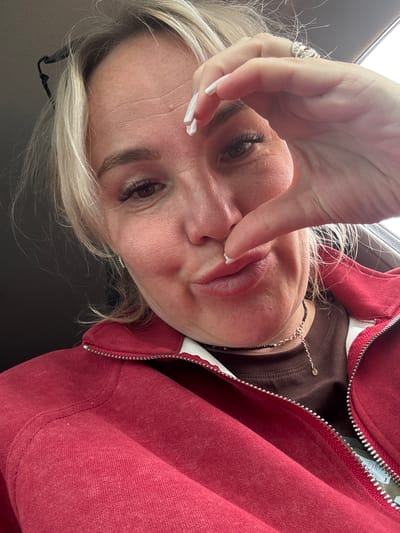 A woman is taking a selfie in a car. She has blonde hair, false eyelashes, and is wearing a red zip-up jacket over a brown shirt and a necklace. Her right hand is raised, with her index finger and thumb making a circle around her eye. She is looking at the camera with her lips pursed. The background is blurred, suggesting the car is in motion. The image was taken on an overcast day. The location is not specified in the image, and there is no information available about Chastyye, Russia.