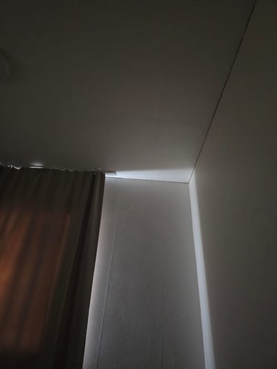 The image shows a corner of a room where the ceiling meets two walls. A curtain hangs on the left side of the frame, obscuring a portion of the wall. The ceiling is textured, with visible seams. A bright light emanates from the junction of the ceiling and the wall, creating a dramatic highlight. The walls are also textured. The scene is dimly lit, with shadows cast by the curtain and the angle of the light. There are no people or animals visible in the image. The location of Chaplynka, Ukraine is not discernible from the image itself.