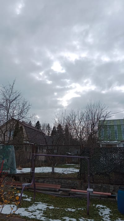 The image depicts a backyard scene in what appears to be the Былина Снт area of Russia. The sky is overcast with grey clouds. In the middle ground are two houses partially visible behind bare trees and fencing. One house has a brown roof and light-colored siding; the other has a green corrugated metal roof. Patches of snow are visible on the ground. A purple metal swing set frame stands in the foreground, with fallen leaves and more patches of snow covering the grass. Yellow flowers are visible in the bottom left corner. A blue barrel is located at the bottom right corner. The fence is a mix of chain-link and some camouflage netting. Some stacked pipes and construction material are visible behind the swing set.