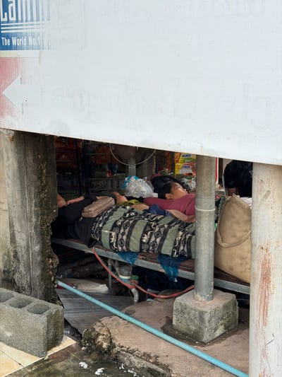 This image depicts a cluttered outdoor scene in Pakxan, Laos, likely a market or roadside stall. A person, possibly female, is partially visible, leaning back and looking upwards. They are wearing a pink top and a patterned skirt or dress. A beige bag is slung over their shoulder, and a smaller clutch bag is held in their hand.

The immediate environment appears unorganized, with various objects piled around. A metal pipe runs horizontally across the frame, supported by what look like concrete blocks and possibly metal scaffolding. Behind the person, shelves are visible, laden with goods, including plastic bottles, bags, and what appears to be food items. A fan is also partially visible in the lower left corner. The ground is a mix of concrete and dirt, with some overgrown vegetation and moss on the concrete surfaces. A light blue pipe runs diagonally across the right side of the image.

The lighting suggests it is daytime, though the specific time is unclear. The overall impression is one of a busy, possibly informal commercial setting. There is some text visible on a white surface in the lower left, which appears to be a sign or advertisement. Part of the text "The World No.1" is legible. The scene conveys a sense of everyday life and commerce in a Southeast Asian setting.