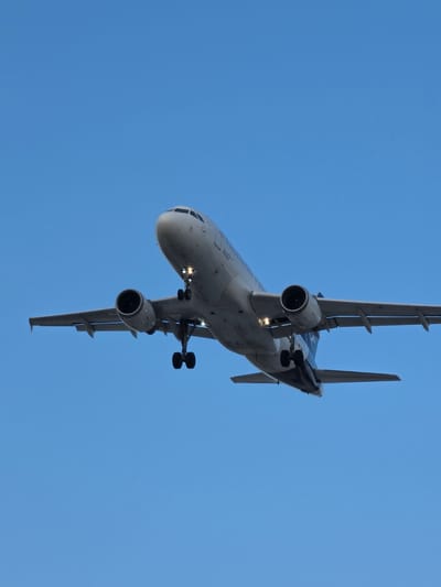A low-angle medium shot captures a commercial airplane ascending against a clear, cloudless blue sky. The aircraft's nose points upward and slightly to the right of the frame, with its landing gear deployed. The underside of the plane reveals two engines mounted under its wings, their landing lights illuminated. The plane's tail section is visible on the right side of the frame, displaying a blue and white logo. The image is taken from a perspective below the aircraft, suggesting the viewer is on the ground looking up. The scene is likely located near Frankfurt Airport in Kelsterbach, Germany, given the airport's proximity to Kelsterbach and the presence of commercial air traffic.