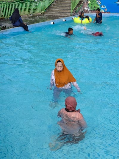The scene takes place at a swimming pool in Kecamatan Tulung, Indonesia, located at the coordinates -7.592 latitude and 110.65 longitude. A woman wearing a hijab, a long-sleeved shirt, and a headscarf is standing in the water in the middle of the pool, facing the camera. Another woman wearing a headscarf is swimming near her, facing away from the camera. A man is swimming towards the left side of the pool. A woman is sitting on a yellow inflatable ring in the pool in the background. A man is standing next to the pool. There is a woman sitting on the side of the pool in the background. The pool is blue and surrounded by a concrete deck. There are stairs leading up to the pool on the right side of the image.