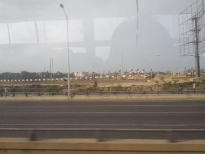 A roadside scene features a multi-lane highway with a barrier. Beyond the barrier, a wide, sandy area hosts numerous flagpoles with flags fluttering atop them. Further back, large, white, domed structures are visible. The sky is overcast, and light poles and an advertising tower punctuate the horizon. The image appears to be taken from inside a moving vehicle, evidenced by window reflections.
