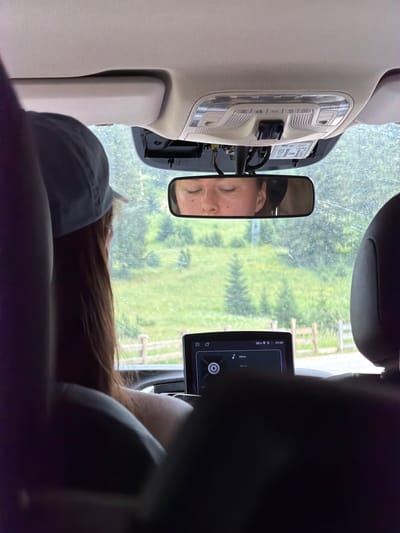 The image shows the interior of a car, looking forward through the windshield. A young woman wearing a light blue baseball cap is visible in the foreground from the shoulders up, facing away from the camera and looking out the window. Her long, brown hair is visible. Her reflection is captured in the rearview mirror, showing her face with her eyes closed and fair skin. The car's dashboard displays a navigation screen showing a time of 09:30. Outside the car, a green, hilly landscape with trees is visible, suggesting a rural or natural environment. There is a wooden fence in the middle ground. The overall scene suggests a journey through a scenic area. There is no information in the image to confirm the location as Trnovo Municipality (FBiH), Bosnia and Herzegovina.