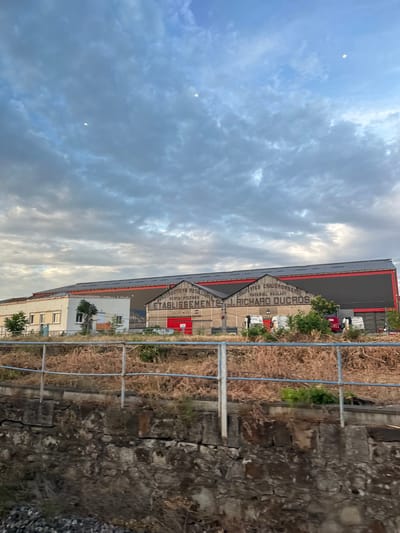 The image depicts a large industrial building complex in the city of Alès, France. The building features a long, dark grey roof with visible solar panels, and a red trim line along its edge. The facade of the building is varied, with sections of lighter colored walls and a prominent section with a gabled roof displaying text. The text on the gabled section reads "CHARPENTES CHAUDRONNERIE", "PONTOS PILONES", "ETABLISSEMENTS J. RICHARD DUCROS", and "MATERIEL ROULANT". Several vehicles, including white vans and a red truck, are parked in front of the building. The foreground shows a dry, overgrown embankment with sparse vegetation and a metal railing. Behind the railing, there is a stone wall and gravel. The sky above is cloudy with patches of blue. There are two small, bright circular lights visible in the sky. The scene appears to be an outdoor industrial setting.