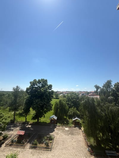 The image depicts a park-like area with trees, green space, and some structures reminiscent of small sheds or playhouses, viewed from a slightly elevated perspective. There are paved areas with a honeycomb pattern, suggesting a walkway or courtyard. The sky is clear blue with a contrail visible. Distant buildings, potentially located in the city of Dulovo, Bulgaria, can be discerned in the background. No people are visible. The general environment appears to be a residential or public green space.