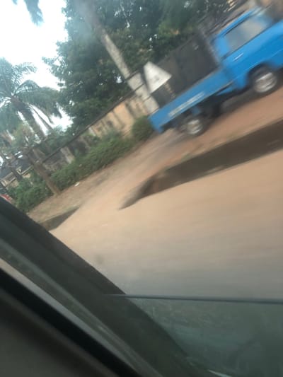 The image captures an outdoor scene, likely viewed from inside a moving vehicle, given the noticeable motion blur and reflections on what appears to be a window in the foreground. The perspective is tilted, with the horizon line running diagonally across the frame.
The dominant subjects are a light blue vehicle, a paved road or path, and lush green foliage. The blue vehicle, which appears to be a van or a small bus, is positioned on the left side of the frame, either parked or moving slowly. It has visible wheels and a boxy shape. White text is discernible on its side; specifically, the word "DANFO" is visible, and below it, another line of text is present but too blurry to read clearly. Near the front of this blue vehicle, there is a white rectangular object or sign with some indistinct dark text on it, possibly "OFO" or "UFO".
To the right of the blue vehicle and the foliage, a light-colored, possibly dirt or unpaved road or path runs roughly parallel. This path appears to be bordered on its far side by a slightly raised edge or curb, separating it from a darker strip of ground or a shallow ditch. Further to the right, the road surface continues, appearing lighter and smoother, possibly paved.
On the far left and in the lower-left corner, dense green trees and vegetation are visible, including the distinctive fronds of a palm tree. In the background, behind the blue vehicle and trees, indistinct shapes of buildings or walls can be faintly seen through the blur.
The image is characterized by significant blur, suggesting either motion during capture or a poorly focused shot. The lighting indicates it is daytime, possibly overcast, as there are no harsh shadows. The reflections in the upper and lower right corners suggest the image was taken through a glass window, with parts of the interior of the capturing vehicle, like a window frame, visible. The overall mood is casual, capturing a moment from transit.