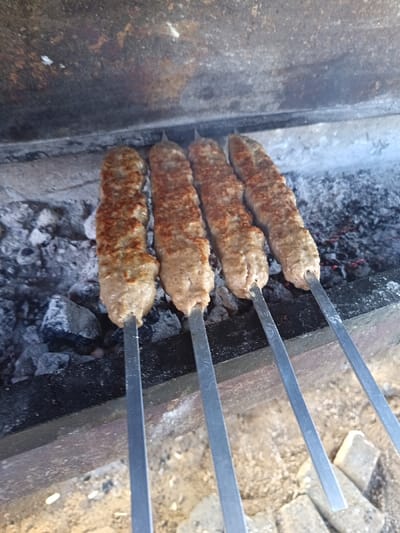 Four skewers of elongated, raw to partially cooked ground meat, likely kebabs, are positioned horizontally over glowing charcoal and ash in a metal brazier. The meat on the skewers exhibits a light brown and pinkish hue, with some areas showing initial browning and reddish seasoning specks. The charcoal beneath is a mix of white ash and black embers. The brazier's interior walls are dark, stained metal, with some lighter concrete or stone elements visible below the cooking area. The ground beneath the brazier appears to be a light-colored, possibly sandy or dusty surface. The scene depicts food preparation outdoors, specifically grilling, in Kasri Arifan, Uzbekistan.