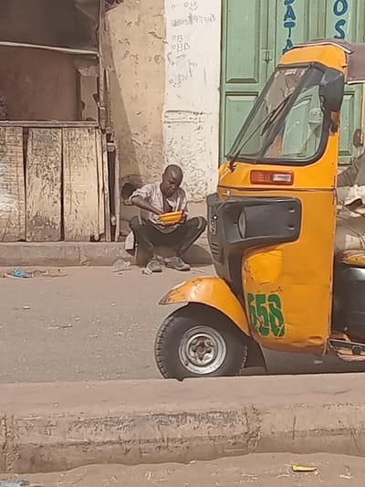 In Dogon Nama, Nigeria, a daylight street scene shows an adult male squatting on the edge of a paved surface. He is wearing a light-colored, short-sleeved collared shirt and dark trousers, with dark shoes visible near his feet. The man holds a bright yellow bowl in his left hand and appears to be scooping food from it with his right hand, his gaze directed at the bowl. To his immediate right, on the paved road, is a yellow three-wheeled motorized vehicle, an auto-rickshaw, with "655" painted in green on its front fender. The vehicle's front windshield, wipers, and the top portion of its cabin are visible, along with its front tire and silver hubcap. In the background, a light-colored, weathered building wall features a section of vertical wooden panels to the left. To the right, green double doors are partially visible, with faint script and partial English words "DATA" and "OS" on the adjacent wall and door. The ground consists of a light-colored paved road and a raised curb, with scattered debris.