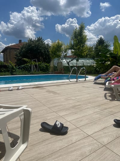 The image depicts a backyard scene with a swimming pool and loungers, located in the city of Madara, Bulgaria. The pool, filled with blue water, is surrounded by a tiled deck. Two pairs of slides, one white and one black Nike slide, are visible on the deck. In the background, a brick house with a terracotta roof and a greenhouse can be seen, along with lush green trees and vegetation under a cloudy blue sky. Two individuals are relaxing on loungers by the pool, their legs visible. The scene suggests a relaxed summer day outdoors.