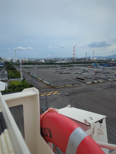 This is an aerial view of a large car storage facility in Yokkaichi, Japan, with an industrial area in the background. The foreground shows the railing of a structure, possibly a ship, with a red life preserver attached. The life preserver has the letters "BOX" visible on it.

The car storage facility is vast, with numerous rows of parked cars, appearing to be new vehicles awaiting transport. The cars are organized in neat lines, with some areas of empty pavement. A road runs through the facility, with a blue truck driving on it. A green grassy area with trees separates the road from a fence.

In the background, a sprawling industrial complex is visible, characterized by numerous smokestacks, some emitting smoke or steam, and various industrial buildings. The sky is overcast with some clouds. The overall scene suggests a port or industrial area, likely involved in the export or import of vehicles and industrial goods.