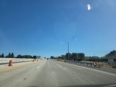 The image depicts a view from a vehicle traveling on a highway. The road is paved with concrete and marked with white lane lines. Orange traffic cones are positioned along the left side of the road, indicating construction or a lane closure. A concrete barrier runs parallel to the road on the left. On the right side, a metal guardrail separates the highway from the shoulder, which is lined with dry grass and some scattered debris. In the distance, trees and vegetation line the sides of the highway, and there are signs for upcoming exits and traffic signals. A few vehicles are visible on the road ahead. The sky is a clear, bright blue, suggesting a sunny day. A light flare is visible in the upper right portion of the sky. The scene appears to be in a suburban or rural area. The overall impression is that of a typical highway commute under clear weather conditions. The time of day is likely midday or early afternoon, given the brightness of the sunlight and the clear sky. There is no visible text that can be clearly read in the image.