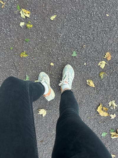 The image shows a top-down view of a person's legs wearing black jeans and light-colored athletic shoes with orange accents. They are standing on a dark, gravelly surface littered with fallen autumn leaves, predominantly yellow and green. The setting appears to be an outdoor path or walkway in the city of Wadern, Germany, characterized by the presence of the worn, asphalt-like ground and scattered leaves, indicative of a park or street environment during the fall season. The person's feet are positioned in a walking stance, with their legs slightly bent at the knees, suggesting movement or a brief pause during a walk. There is no visible interaction between the person and any other individuals or objects in the immediate vicinity. The lighting suggests an overcast day, typical of autumn weather.
