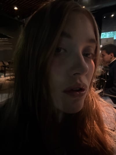 A close-up, low-lit portrait shows a fair-skinned person with red hair. The face, angled towards the camera, features prominent nose and lips. A second individual is visible in the background, partially obscured. The overall image has a casual, candid feel, possibly taken indoors. The limited lighting casts shadows that obscure details.