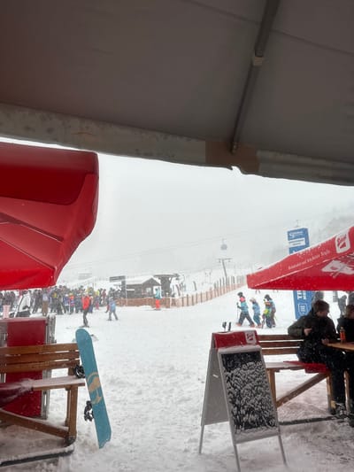 The image depicts a snowy winter scene at a ski resort, specifically within the Gastein ski region, including Bad Hofgastein, Austria, as indicated by visible signage. The perspective is from beneath a light-colored tent or canopy structure, which partially covers the upper portion of the frame. A heavy snowfall is ongoing, with visible flakes in the air and snow accumulation on surfaces.
In the foreground, to the left, a portion of a red sun umbrella is visible. To the right, another red sun umbrella, branded with "Stiegl" and the text "Redlichkeit auf höchster Stufe," shades a wooden picnic-style table and bench. Two individuals are seated at this table; one, wearing a dark jacket, is visible from the back. A light blue snowboard with black bindings is propped vertically against a wooden structure in the left-center foreground. An A-frame sign, black with a white frame, stands in the center-right foreground, its surface heavily covered in fresh snow, rendering its text illegible.
The midground reveals a wide, snow-covered ski slope populated by numerous skiers and snowboarders. On the far left, a large group of individuals in ski gear is gathered, possibly in a queue or waiting area. Scattered across the slope are other skiers, including one in an orange jacket and a group of three younger individuals in colorful ski suits (blue, yellow, green) walking with ski poles. Several wooden structures, possibly huts or service points, are visible on the slope, with hay bales surrounding some. Ski lift poles and cables extend up the slope into the background, and a gondola or chairlift cabin is faintly discernible in the upper midground. Blue signs with "Gastein," "SPORTGASTEIN," and "Bergbahnen GASTEIN" are visible in the background, confirming the resort location. The environment is overcast and appears cold.