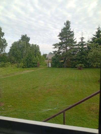 The image depicts a backyard scene in the village of Maļutki, Latvia, on a cloudy day. The foreground is dominated by a neatly mowed green lawn. A brown metal railing runs diagonally across the bottom of the frame, suggesting a vantage point from a porch or deck. Further into the yard, a dirt path leads towards a gate, which is partially open. Beyond the path, a dense line of trees and bushes forms a natural boundary, with a large pine tree standing prominently on the right side of the frame. In the distance, a few houses with light-colored roofs are visible through the trees. The sky is overcast with a pattern of layered clouds.
