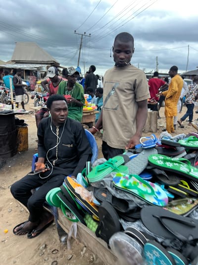 In Oko Amakom, Nigeria, under an overcast sky, a street market scene unfolds. Two men are the primary subjects. In the foreground, seated on a blue plastic chair, a man in a black traditional outfit with white earphones is holding a smartphone and looking to his right. Beside him, a large display of colorful flip-flops and sandals is arranged on a wooden structure. Behind him, standing and looking forward, is another man wearing a tan t-shirt. A bustling market atmosphere is suggested by the presence of other individuals in the background, motorcycles, and makeshift stalls with corrugated metal roofs. The ground is dirt and scattered with small stones.