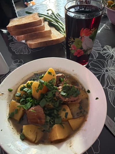 A high-angle, close-up shot shows a table setting with a meal and a drink. In the foreground, a white plate holds a stew with large chunks of meat and potatoes, garnished with chopped green herbs. To the left of the plate, several slices of toasted bread are stacked, next to a bunch of green beans. A tall glass with a dark red beverage, possibly wine, is positioned behind the food, decorated with a colorful bird and floral design. The table appears to be covered with a dark tablecloth featuring a floral pattern. The setting suggests a meal is being served or enjoyed. The lighting indicates it could be daytime or an indoor setting with artificial light. There is no visible text in the image. The location is identified as Krasnyi Krym, Russia.
