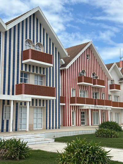 The image displays a row of three multi-story buildings, characteristic of the "palheiros" architectural style, located in Costa Nova do Prado, within the municipality of Ílhavo, Portugal. The building on the far left features a facade clad in alternating vertical blue and white stripes. The central building is similarly adorned with alternating vertical red and white stripes. A third building to the right is partially visible, featuring a predominantly white facade with a brown-tiled roof. All buildings are topped with pointed gable roofs. Each structure incorporates multiple white-framed windows, some with arched upper sections, and balconies with white undersides and terracotta-colored, geometrically patterned railings. The ground level of the blue and white striped building includes recessed entrances supported by white columns. In the foreground, a neatly manicured lawn with several clusters of broad-leafed green plants (Agapanthus spp.) is visible, traversed by light-colored paved walkways and steps leading towards the buildings. The scene is set under a clear blue sky with scattered wispy white clouds. No individuals, vehicles, or discernible actions are present in the image.