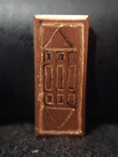 The image shows a close-up, eye-level view of a small, rectangular, copper-colored object with an engraved design. The design appears to be a stylized depiction of a building, possibly a house or a temple, with a triangular roof and multiple rectangular openings. The object is oriented vertically and has a metallic sheen. The background is dark and out of focus, with a slightly textured surface in the foreground that is also out of focus. The object is centrally framed in the image. No specific location within Sokolohirsk, Ukraine, can be determined from this image.