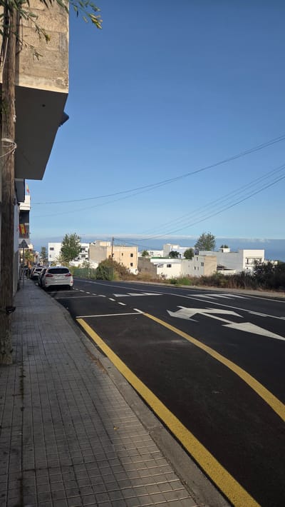 This is a daytime, exterior, eye-level, full-frame, vertical photograph taken in Guía de Isora, Spain, likely on a road leading towards the coast. The foreground features a paved sidewalk with square grey tiles, bordered by a curb and a paved road with fresh asphalt marked with double yellow lines and white directional arrows. To the left, a portion of a building is visible, with a beige, rough-textured exterior and a cantilevered overhang. A wooden pole with white electrical wires attached stands near the building.
The middle ground displays a row of parked cars along the sidewalk, including a silver hatchback. Beyond the cars, there is a grassy embankment and a line of low-rise buildings with white and beige facades. Trees and other vegetation are scattered between the buildings.
The background consists of a clear, bright blue sky with a few wispy clouds near the horizon. A faint line of what appears to be the ocean or a body of water is visible in the distance, under the horizon line. Overhead, several electrical wires stretch across the sky. Two Spanish flags are visible hanging from balconies on the building on the far left. The overall scene depicts a quiet street in a residential or commercial area, possibly during a sunny afternoon.