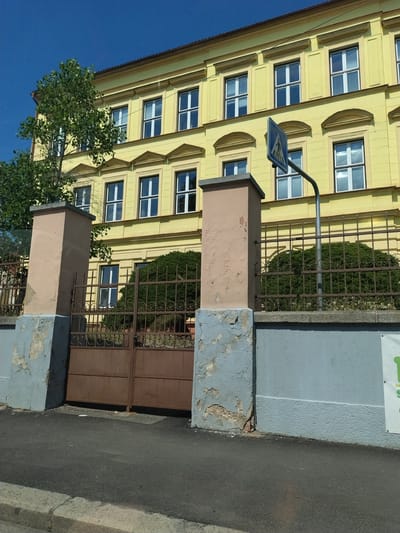 The image captures a sunny daytime scene in front of a yellow building, likely a school or public institution, located in Horažďovice, Czechia. The building exhibits classical architectural elements with multiple rows of evenly spaced windows, some featuring decorative arched pediments. A tall, mature tree with lush green foliage stands to the left of the building, partially obscuring some of its windows and architecture.
In the foreground, a brown metal gate with ornate scrollwork is visible, flanked by two concrete pillars. The pillars have a light peach-colored surface layer that is peeling and cracked in places, revealing a grey underlayer and brickwork. A low grey concrete wall runs along the perimeter of the property. A pedestrian crossing sign is mounted on a metal pole to the right of the gate. Behind the gate, a dense, well-maintained green hedge is visible, suggesting a manicured garden or courtyard.
The ground in front of the gate is paved with asphalt, bordered by a concrete curb. The sky is a clear, bright blue, indicating fair weather. The overall impression is one of an established, somewhat aged public building set against a backdrop of clear skies and greenery. There are no people visible in the image.
