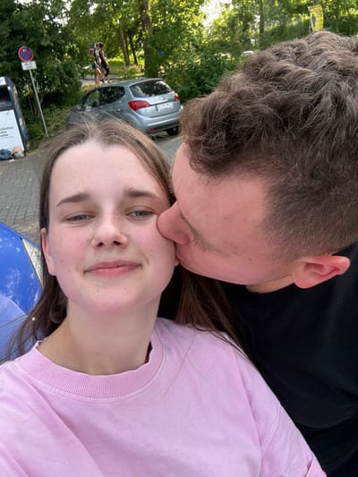 This is a selfie of a young couple outdoors, with a man kissing a woman on the cheek. The man has short, brown, curly hair and is wearing a black t-shirt. The woman has long, brown hair and is wearing a pink t-shirt. She has freckles on her face and is smiling at the camera. In the background, there is a silver car, trees, and a street sign. The time of day appears to be daytime, and the weather seems to be warm and sunny. There is some text visible on the street sign and on a poster in the background, but it is mostly illegible. The overall mood of the image is romantic and happy.