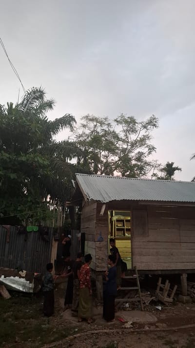 In Grong-grong, Indonesia, a wooden house with a corrugated metal roof stands on stilts. The house is elevated, and a wooden staircase leads to an open doorway. Inside, shelves are visible. Several young males, some wearing traditional Islamic attire including peci hats and sarongs, are gathered outside the house. Some are interacting with each other, while others appear to be entering or exiting the structure. The surrounding environment is rural, with lush vegetation including palm trees visible to the left and in the background. A corrugated metal fence or wall is present to the left of the house. The sky is overcast, suggesting it may be early morning or late afternoon.