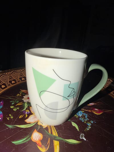 This media file features a white ceramic mug with a light green handle, decorated with abstract black lines and light green geometric shapes. The mug is positioned at an angle, resting on a dark brown tray. The tray is adorned with a floral pattern, including stylized flowers in shades of orange, yellow, and white, interspersed with green leaves and blue blossoms. Small hearts are visible within a decorative border at the bottom of the tray. The background is dark and out of focus, suggesting a dimly lit indoor setting. No people or specific activities are depicted. The overall presentation highlights the mug and its decorative tray. There is no visible text in the media file.