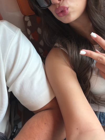 A close-up, high-angle shot captures a person resting or relaxing in what appears to be an indoor setting or vehicle in Cagnes-sur-Mer, France. The primary subject is the upper body of an individual, with long, dark brown hair spread across the frame. The left side of their face is visible, featuring partially pursed lips with dark lipstick and a mole on the chin area. Dark sunglasses rest near their face. One hand, adorned with white-painted square-tipped fingernails, is visible near the hair.
The person wears a white long-sleeved top, likely a sweatshirt or t-shirt, and a blue denim garment can be seen on the right side of the frame. A dark mole is also visible on their right arm. Underneath the person's head and hair, a patterned fabric with brown/orange and white/grey abstract shapes suggests upholstery or a blanket, reinforcing the impression of a comfortable, relaxed environment, possibly in transit or at home. No specific activity beyond resting is clearly depicted, and no text is visible. The time of day is not discernible from the indoor lighting.