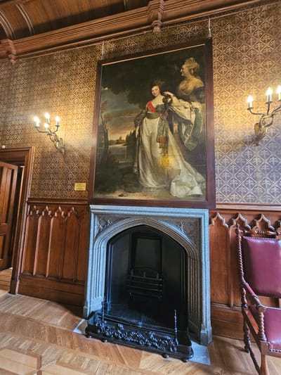 The image shows a room interior. On the wall is a framed painting of a woman in a light-colored dress being attended by another person. The wall has patterned wallpaper. Below the painting is a stone fireplace. To the right of the fireplace are wooden panels and a chair with a maroon cushion. Above the scene, hanging from the ceiling, is a chandelier with multiple light bulbs. The location is likely a building interior, but specific details that would place it within Алупкинский городской совет, Ukraine are not evident from the image itself.