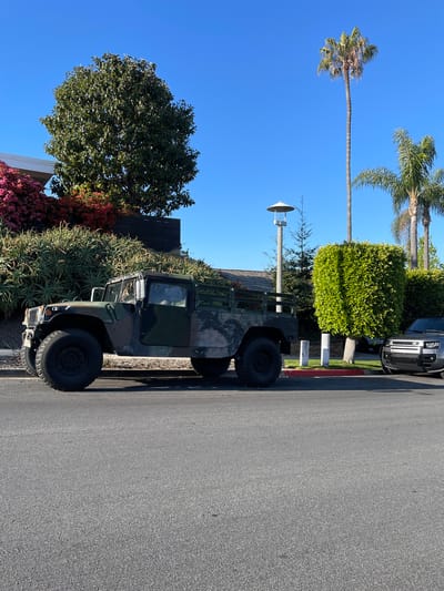The image depicts a Humvee military vehicle parked on a street in Newport Beach, California. The vehicle is positioned parallel to the curb, near landscaping that includes succulents, trees, and bushes with red and pink flowers. Further down the street, there's a manicured bush and a modern black SUV. Palm trees are visible in the background against a clear blue sky. A street lamp stands nearby, contributing to the residential atmosphere. The scene suggests a sunny day in an affluent residential area.
