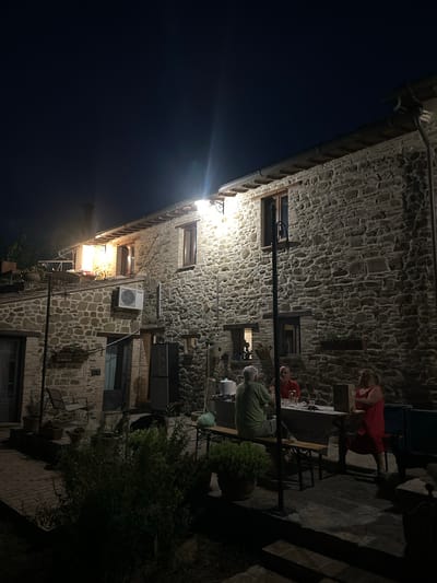 The image depicts a stone building at night, with warm light emanating from windows on the upper floors. Several people are gathered around a table in the foreground, seated on a wooden bench. The table is set with plates, glasses, and a bottle. The building's exterior is constructed of rough-hewn stones, and an air conditioning unit is visible on the wall. The ground around the building appears to be paved with bricks and concrete. A dark-colored dog is present near the table. The scene suggests a social gathering taking place outdoors on a warm evening. The location is identified as Cannara, Italy.